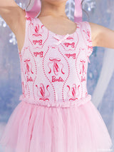 A young girl wearing a pink sleeveless leotard top with pink ballet shoe prints and 'Barbie' logos, paired with a matching pink tulle tutu skirt, shown against a soft icy blue background from Tutu du Monde x Barbie colleboration.