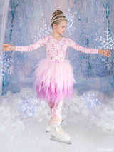 A young girl in a Barbie pink winter ballet costume with a fluffy tutu skirt and ice skates, posing in a snowy, icy-themed studio setting.